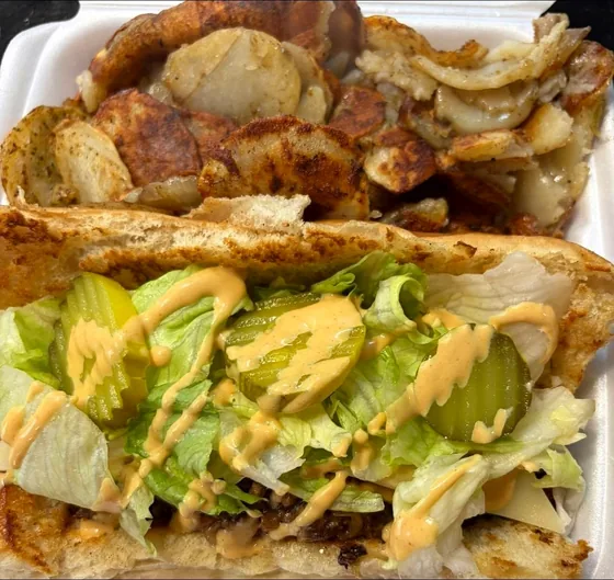 Philly cheesesteak on a toasted hoagie with lettuce, pickles and sauce, next to seasoned fried potatoes